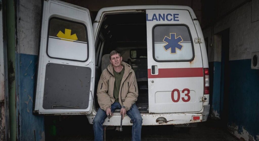 На фото задні двері автомобіля швидкої допомоги з цифрами 03 червоним кольором на білому фоні і червоною смужкою горизонтальною. Одна половинка дверей відчинена і на бампер сів русявий чоловік у джинсах, світло-коричневій куртці. , який у руках тримає металеві предмети, схожі на гайковий ключ та запчастину.