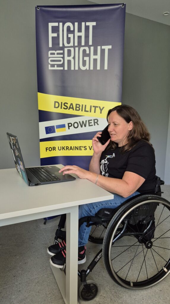 Наталя Шишолік, голова громадської організації «Осіб з інвалідністю “Довіра”» на кріслі колісному за столом з ноутбуком (laptop) і працює. На фоні стоїть банер організації Fight For Right. Наталя - молода жінка з темно-русявим волоссям. Вона одягнена в Чорну футболку з білим контурним малюнком спереду, сині джинси та чорні кросівки з оранжевими шнурівками