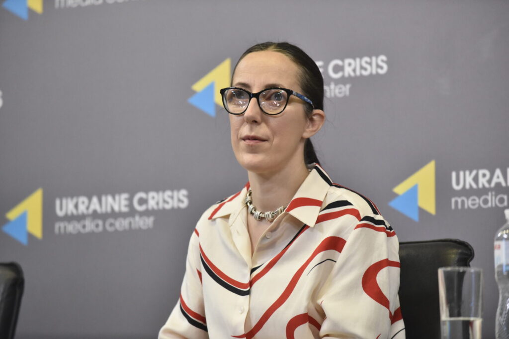 Фото Юлії Сачук. Юлія – молода жінка з темним волоссям, зібраним у хвіст. У неї окуляри в темній оправі. Юлія одягнена в світлу сорочку з хвилястими смугами червоного та чорного кольорів. На фоні банер з сірим тлом, на якому написано Український кризовий медіацентр і його логотип з трикутниками, накладеним один на одного жовтого та блакитного кольорів