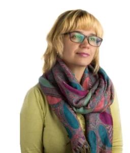 На фото Ірина Трохим. Світловолоса жінка в окулярах. Одягнена в зелену кофтину, навколо шиї накинутий різнобарвний шарф темного кольору.