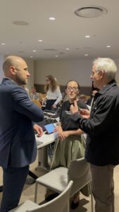 Юлія Сачук стоїть по центру між двома чоловіками в темному костюмі один, інший у чорній куртці. На Юлії чорна футболка і спідниця кольору хакі. У Юлії окуляри в чорній стильній оправі. Волосся зібране у хвіст.