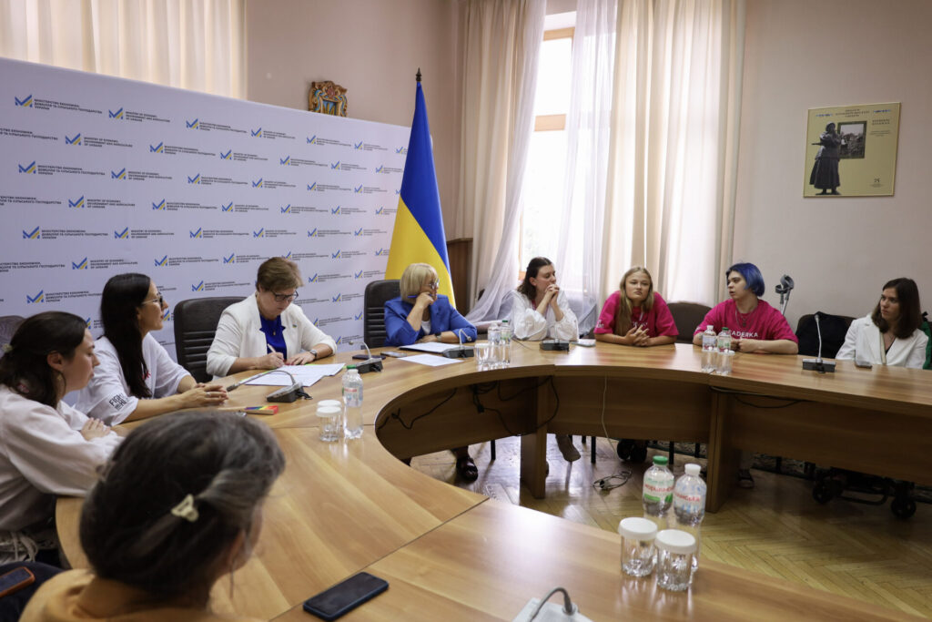 За овальним світлокоричневим столом сидять учасниці та організаційна команда Fight For Right разом з Катериною Левченко, урядовою уповноваженою з питань гендерної політики (шатенка з короткою стрижкою, яка сидить лівіше від центру в білому костюмі і синій блузі, в окулярах, робить нотатки ручкою на аркушах перед собою) та її заступницею пані Галиною (світловолоса жінка в синьому жакеті на фоні прапора України). Праворуч від пані Галини три учасниці Школи Лідерка та асистентка проєктів крайня в білому жакетів Олександра Вовченко. Лівіше від Катерини Левченко Юлія Сачук (брюнетка з довгим волоссям у білій футболці, з надписом на рукаві Fight For Right), учасниця Школи Діана Палко (брюнетка з волоссям до плечей, одягнена у білу блузу) та на передньому фоні спиною видно голову з зачіскою та плечі в жакеті карамельного кольору Яни Салахової, фасилітаторки Школи. На фоні лівіше банер з логотипом одного з міністерств, оскільки захід відбувавася в Кабінеті Міністрів України. Правіше від прапора на фоні вікно зі світлими фіранками, пастельні стіни бежевого кольору, на яких подекуди висять картини з портретами видатних українців та українок та короткою інформацією про них.