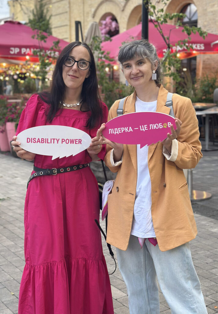 На фото зліва Юлія Сачук (темноволоса жінка в окулярах, яскравій сукні кольору "фуксія", з намистом на шиї та чорним ременем на поясі з металевими округлими вставками). Поруч з нею Яна Салахова (жінка з сивим волоссям з чубчиком, одягнена в блакитні джинси, білу футболку з жакетом карамельного кольору поверх неї, з рюкзаком та оригінальними сережками світлого кольору). Юлія тримає в руках табличку овальної форми для фотосетів "Disability Power", а Яна - "Лідерка – це любов". На фоні споруда жовтого кольору, де відбувався випускний у лідерок, парасольки рожеві зі столиками та стільцями біля ресторацій і багато яскравих квітів у вазонах. 